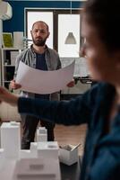 Standing architect holding blueprints thinking about architectural design working together with colleague engineer in modern office. Architects doing creative teamwork on 3d buildings maquette. photo
