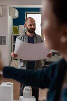 Standing bearded architect looking focused at construction plans collaborating with colleague engineer in modern office. Architects doing architectural design creative teamwork on foam scale model. photo