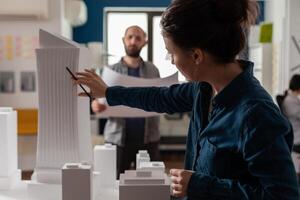 Architect inspecting futuristic building design of skyscraper with colleague engineer holding blueprints. Architects doing teamwork at desk with white foam maquette of construction project. photo