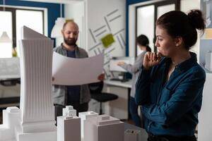 Architect in modern office thinking about design work standing next to tall building foam model with team colleague engineer holding blueprints. Team of architects doing creative thinking. photo