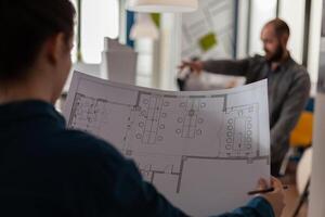 Over shoulder view of architect holding blueprints in architectural office doing teamwork with collegue engineer in the background. Closeup of construction plans of modern business floor plan. photo