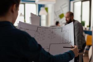 Closeup of architect in architectural office holding pencil and blueprints with floor plan of business building. Over shoulder detailed view of construction plans of modern design real estate project. photo