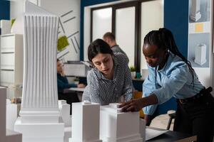 Architectural engineers doing teamwork reading construction plans over table with white foam scale model of buildings. Team of two architects colleagues collaborating looking down at blueprints. photo