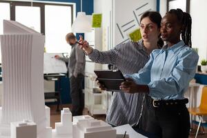 Woman architect pointing pencil at skyscraper architectural model standing next to colleague engineer holding tablet in modern office. Architects doing teamwork looking at residential project maquette. photo