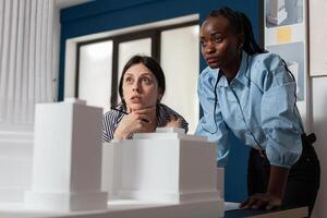 Project engineers women colleagues looking inspired in front of 3d residential maquette in architectural modern office. Focused professional architects team inspecting skyscraper white foam model. photo