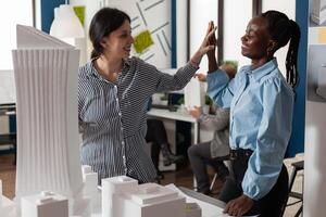 Team of two women architects high fiving standing in front of table with foam scale model of residential buildings in architectural modern office. Successful happy engineers celebrating team project. photo