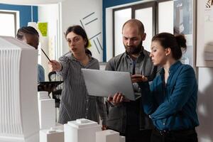 Couple of professional architects holding laptop looking at blueprints next to colleagues with tablet pointing at 3d maquette of skyscraper. Team of architectural engineers doing creative teamwork . photo