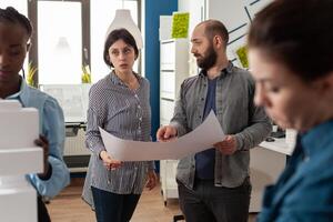 Architects couple holding blueprints in modern architectural office explaining design process to team of colleagues. Creative team reading construction plans for real estate urban developing. photo