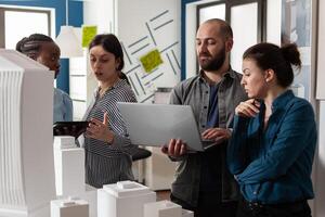Team of professional architects meeting at office analyzing blueprints on laptop computer and digital tablet. Architectural engineers doing teamwork next to maquette building model project. photo