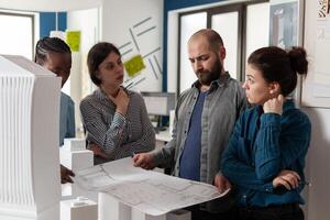 Team of engineers looking at architect holding blueprints for construction project in modern architectural office. Professional engineers analyzing paper plans for building in front of foam maquette. photo