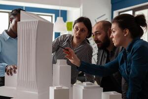 Professional architect with working partners pointing at skyscraper foam scale model in architectural office. Team of engineers collaborating on group project at desk with buildings maquette. photo