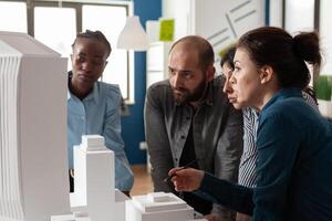 Professional architects discussing architecture design of skyscraper foam scale maquette in architectural office. Team of engineers working together on group project at desk with buildings model. photo