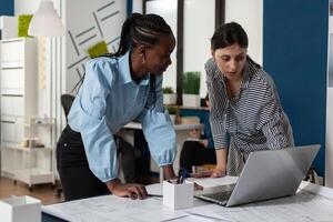 Team of two professional women architects working on laptop computer with construction plans on desk in architectural office. Engineers designing blueprints for real estate project. photo