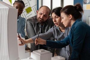 Two women professional architects explaining construction model design to team colleagues in front of white foam maquette. Engineers working together on residential project in architectural office. photo