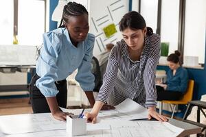 Focused team of women architects inspecting construction plans standing at desk in modern architectural office. Team of two engineers colleagues discussing blueprints for residential project. photo