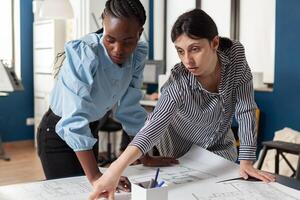 Dedicated team of engineers colleagues discussing construction plans standing at desk in modern architectural office. Team of two women architects discussing blueprints for urban development project. photo