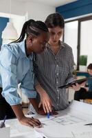 Architect holding digital tablet computer with construction plans explaining blueprints to african american engineer colleague. Professional architects in architectural office designing urban development project. photo