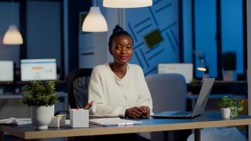 Portrait of business black woman looking at camera smiling raising head from laptop working in start-up financial company late at night. Focused employee doing overtime respecting deadline of project photo