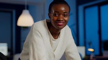 Close up of business african woman smiling at camera after typing on laptop working in startup financial company late at night. Focused employee doing overtime for job respecting deadline of project photo