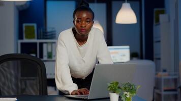 African business woman standing near desk looking at camera after reading mails on laptop working in start-up company late at night. Focused employee using technology network wireless doing overtime photo