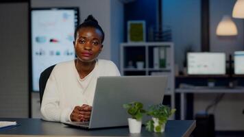 Exhausted dark skin businesswoman typing an email late at night and looking in camera sighing working late at night in start-up business office. Focused employee doing overtime for job respecting deadline photo