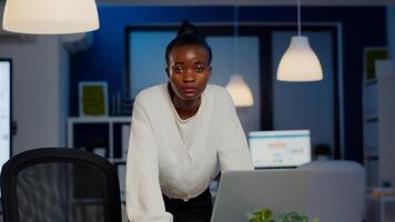 Focused tired business woman looking at camera after reading tasks on laptop standing near desk in start-up company late at night. Employee doing overtime for job respecting deadline photo