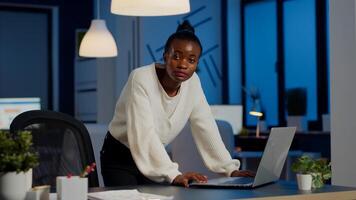 Manager black woman standing at desk searching on laptop, typing and looking tired at camera in start-up company. Focused employee reading tasks doing overtime for job respecting deadline of project photo