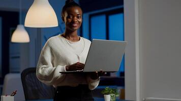 Black business woman looking at camera smiling holding laptop standing near desk in start-up company late at night. Focused employee using technology network wireless doing overtime for job photo