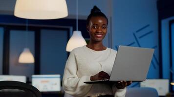 Happy african freelancer looking at camera smiling holding laptop standing near desk in start-up company late at night. Focused employee doing overtime for job respecting deadline of financial project photo