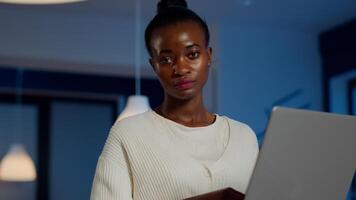 Tired african manager looking at camera sighing holding laptop typing on it, working late at night in start-up business office. Focused employee doing overtime for job respecting deadline of project photo