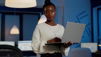 Exhausted black businesswoman holding laptop typing and sighing looking at camera standing in start-up company late at night. Focused employee doing overtime for job respecting deadline of project photo