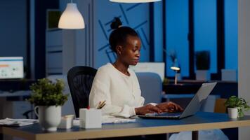 Busy african manager working on financial reports checking statistics graphs, typing on laptop sitting at desk late at night in start-up office doing overtime to respect deadline of financial project photo
