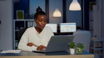 Stressed african manager woman working with financial graphs checking graphs, typing on laptop, reading raports late at night in start-up office doing overtime to respect deadline of project. photo
