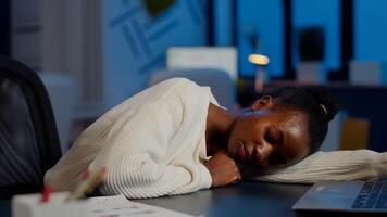 Dark skin freelancer doing overtime falling asleep with hand on desk trying to respect deadline for project. African employee working late at financial project sleeping on table in strat up office photo