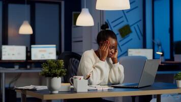 Overworked african manager woman massaging head while sitting at workplace in start-up office working overtime at laptop. Exhausted tensed businesswoman respecting deadline coping with migraine photo