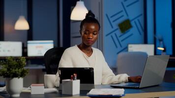 Multitasking black business woman working at laptop and tablet in same time doing overtime in start-up office. Busy african employee analysing financial statistics overworking writing, searching. photo