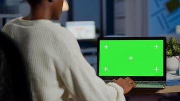 African woman manager looking at mock up monitor of laptop, green screen mockup, chroma key desktop sitting at desk in business office late at night. Freelancer watching at isolated desktop overwork photo