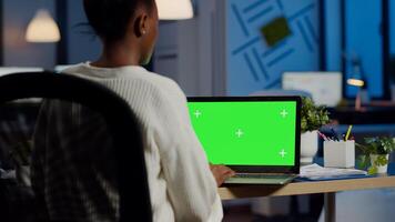 African businesswoman reading on laptop with greenscreen display, green mockup, chroma key monitor sitting at desk in business office late at night. Freelancer watching at isolated desktop overwork photo