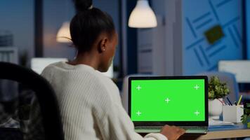 Dark skin freelancer working in front of greenscreen display of laptop sitting at desk in business office late at night. Businesswoman watching desktop with green mockup, chroma key, working overtime photo