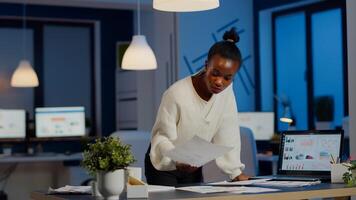 Stressed african manager woman working with financial documents standing at desk checking graphs, holding papers, reading raports late at night in start-up office doing overtime to respect deadline. photo