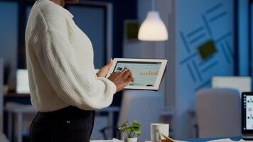 African freelancer woman holding tablet checking financial graphs standing in start-up office late at night. Businesswoman using social network, texting and blogging working overtime for job photo