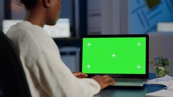 Black freelancer searching using laptop with greenscreen display, green mockup, chroma key desktop sitting at desk in business office working at night. Freelancer watching at isolated desktop overwork photo