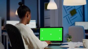 Back view of black business woman looking at laptop with greenscreen, green mockup, chroma key desktop sitting at desk in start up business office overworking. Freelancer watching at isolated display photo