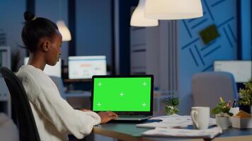 African manager typing at laptop with chroma key display during night time in start up business office working overtime. Freelancer watching mockup desktop monitor with green screen, writing datas photo