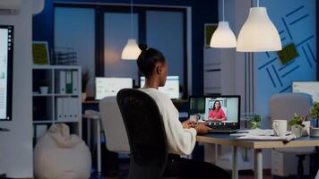 African freelancer working remotely discussing with woman partner online sitting in front of laptop in start-up office talking during virtual meeting at midnight, using headphone doing overtime photo