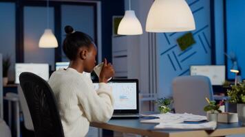 Busy african business woman analysing financial reports checking company statistics graphs, looking at laptop, pointing to numbers late at night in start-up office doing overtime to respect deadline photo
