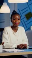 Portrait of business black woman looking at camera smiling raising head from laptop working in start-up financial company late at night. Focused employee doing overtime respecting deadline of project photo