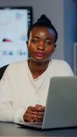 Exhausted dark skin businesswoman typing an email late at night and looking in camera sighing working late at night in start-up business office. Focused employee doing overtime for job respecting deadline photo