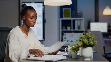 Black manager analysing graphics and financial statistics from documents working overtime in front of laptop sitting in start-up business office. Focused employee respecting deadline of project photo
