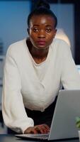 African business woman standing near desk looking at camera after reading mails on laptop working in start-up company late at night. Focused employee using technology network wireless doing overtime photo